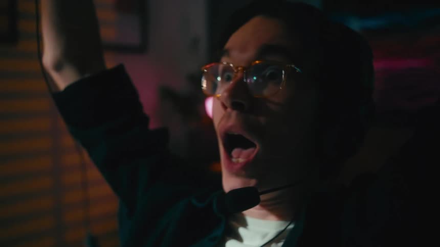An emotional gamer with glasses screams with excitement while playing on a console with a controller in a dimly lit room