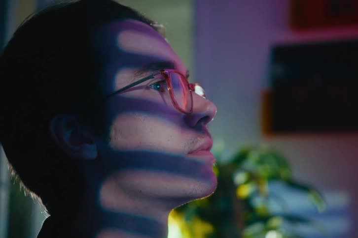 Artistic close-up profile of a young man in glasses looking up with colorful light and shadows projected on his face