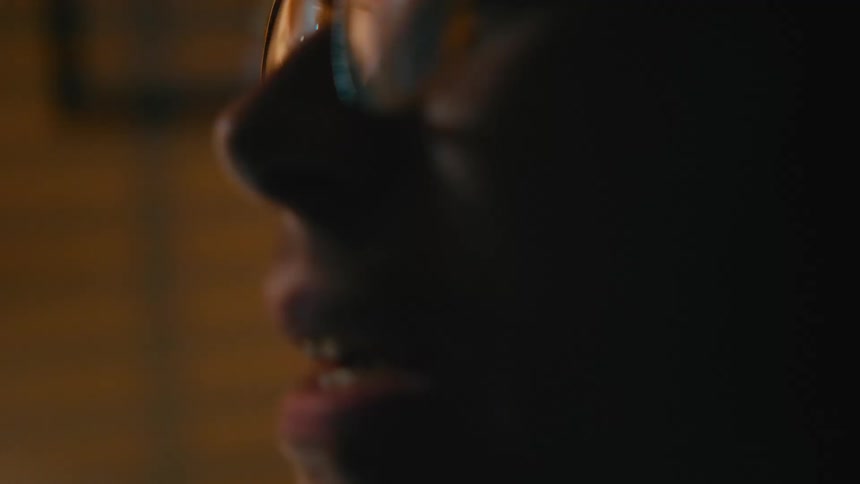 A shaky, out-of-focus close-up profile of a man in glasses talking animatedly in a dimly lit, warm environment