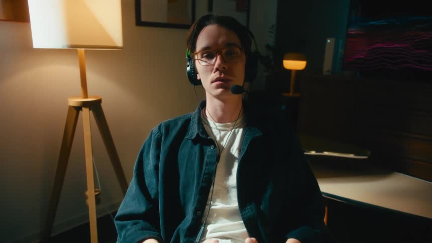 A young man with glasses and a headset sits intently focused on a video game, holding a controller in his dimly lit room at night