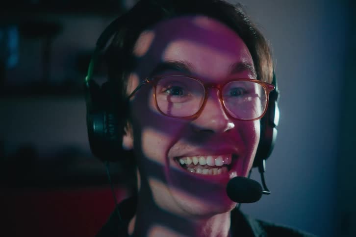 An overjoyed gamer wearing a headset and glasses smiles intensely at his screen in a neon-lit room