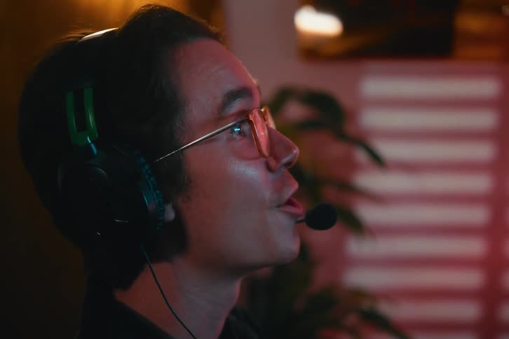 Close up profile of a young man with glasses and a headset talking and smiling while playing video games online