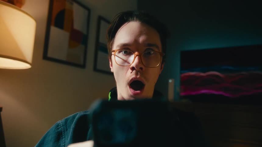Dramatic vertigo zoom effect on a shocked young man with glasses staring at his smartphone screen in his apartment at night