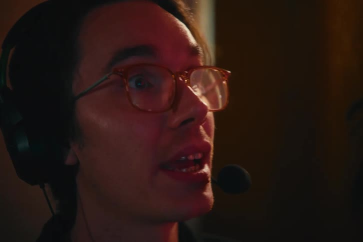 A focused young man in glasses wearing a gaming headset talks excitedly while playing a video game in a dimly lit room