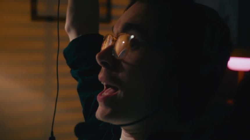 An excited gamer wearing glasses and a headset shouts in victory while playing video games in his dimly lit room