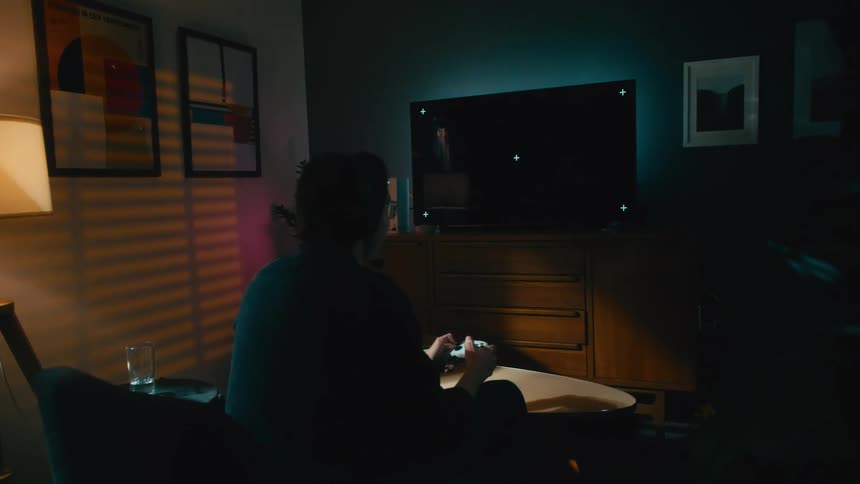 A person wearing headphones plays a video game on a TV with tracking markers in a dark, stylish living room