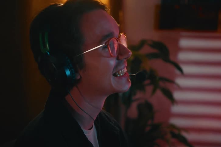 A young man with glasses and a headset rubs his tired eyes in frustration before erupting in triumphant laughter while playing video games