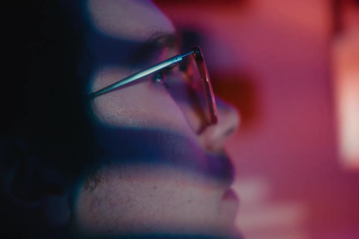 Close-up profile of a young man with glasses engrossed in watching a screen with vibrant neon lights reflecting on his face