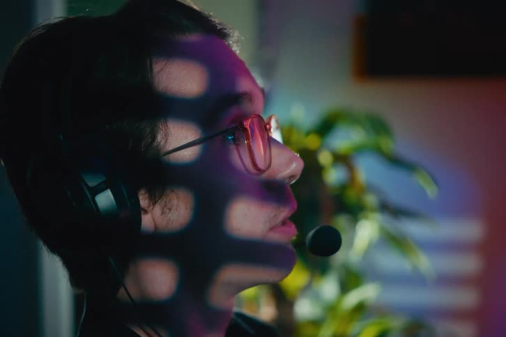A focused young man with glasses and a gaming headset talks into his microphone with intense concentration, his face cast in shadows and neon light