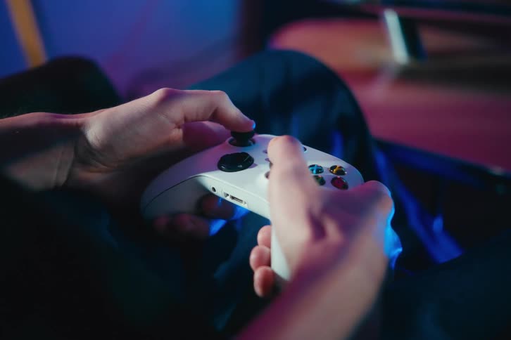 A gamer's hands are shown in close-up as they use a white controller to play a video game in a neon-lit room