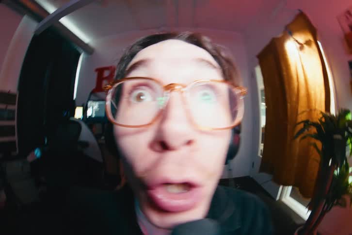 An energetic male streamer with glasses and a headset makes comical and surprised expressions into a fisheye camera