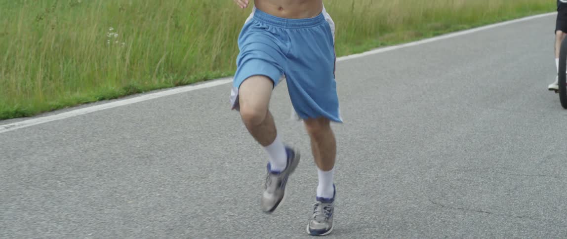 Shirtless man jogging on pavement as another man on a bike rides behind
