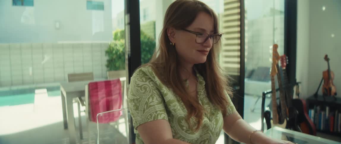 A young woman with glasses smiles while working on her laptop at a glass table in a sunny, modern home office as the camera pushes in
