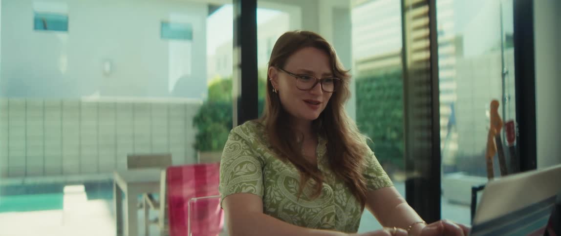 A woman working on her laptop in a sunlit modern home office smiles and then laughs with joy as the camera slowly zooms in