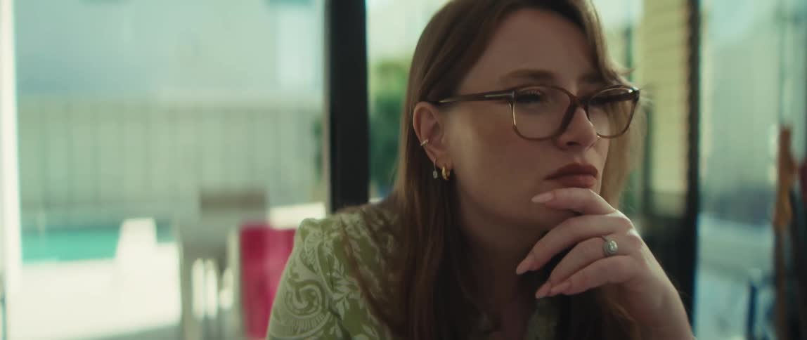 A thoughtful young woman with glasses sits in a sunlit modern room, deeply contemplating with a serious expression