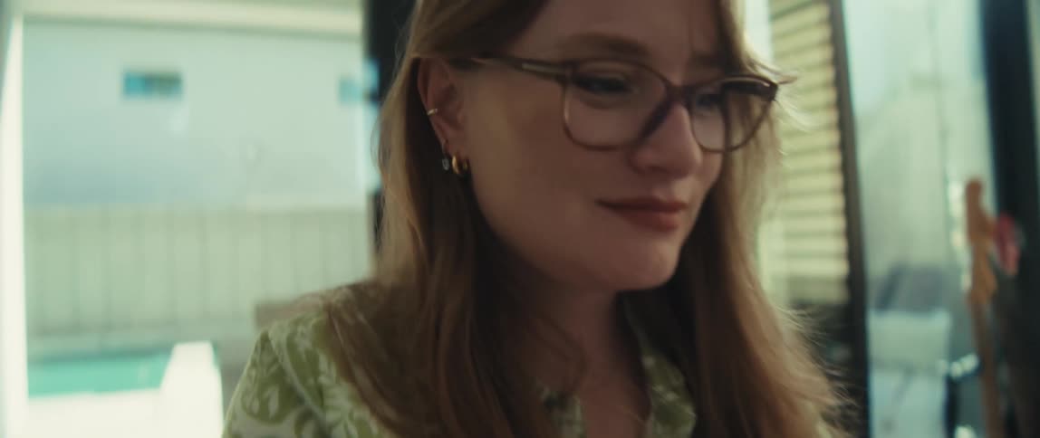 A young woman with red hair and glasses smiles as she types on her laptop in a modern setting