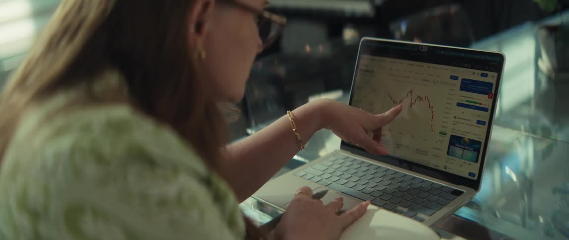 A woman with glasses works on her laptop, pointing at and analyzing a financial chart showing market trends
