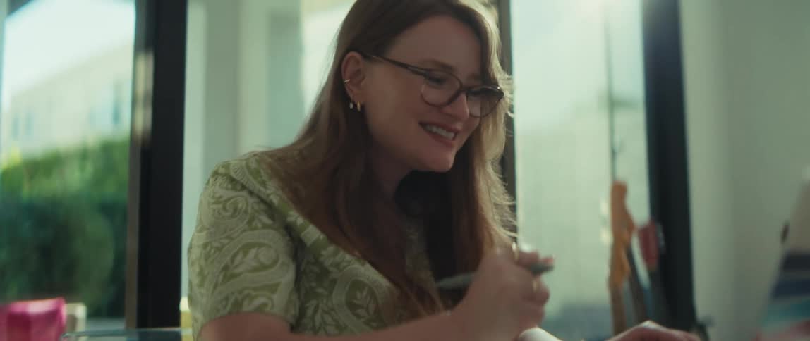 A focused young woman wearing glasses works on her laptop in a bright room, her expression changing from concentration to a happy smile as she types