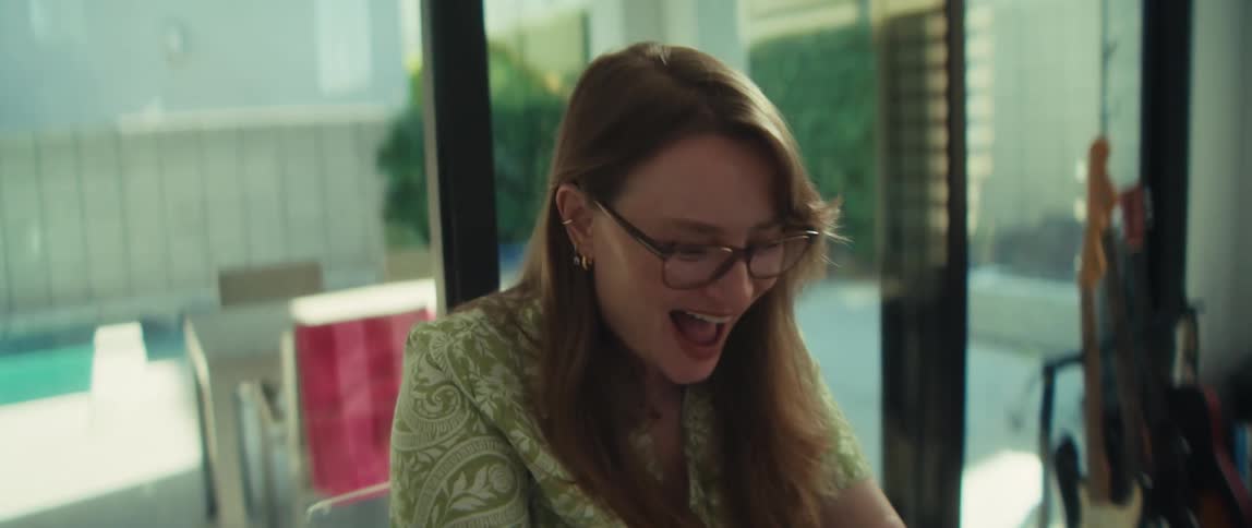 An overjoyed woman with glasses laughs while looking at her laptop screen in a modern home with a pool in the background
