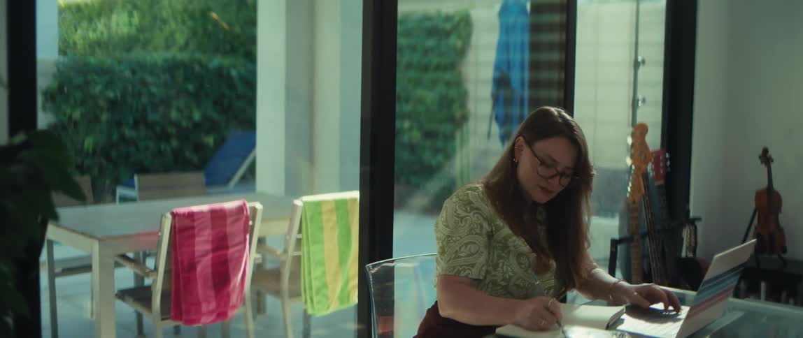 A woman with glasses works from her modern home, taking notes in a journal while looking at her laptop