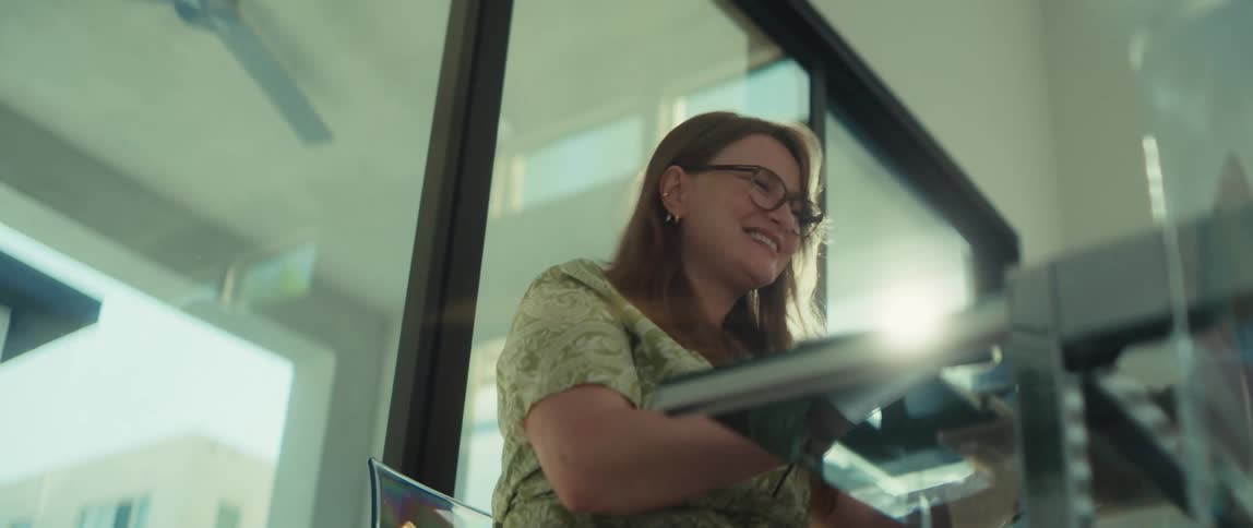 A cheerful woman with glasses laughs heartily while sitting at her desk in a bright, sunlit office