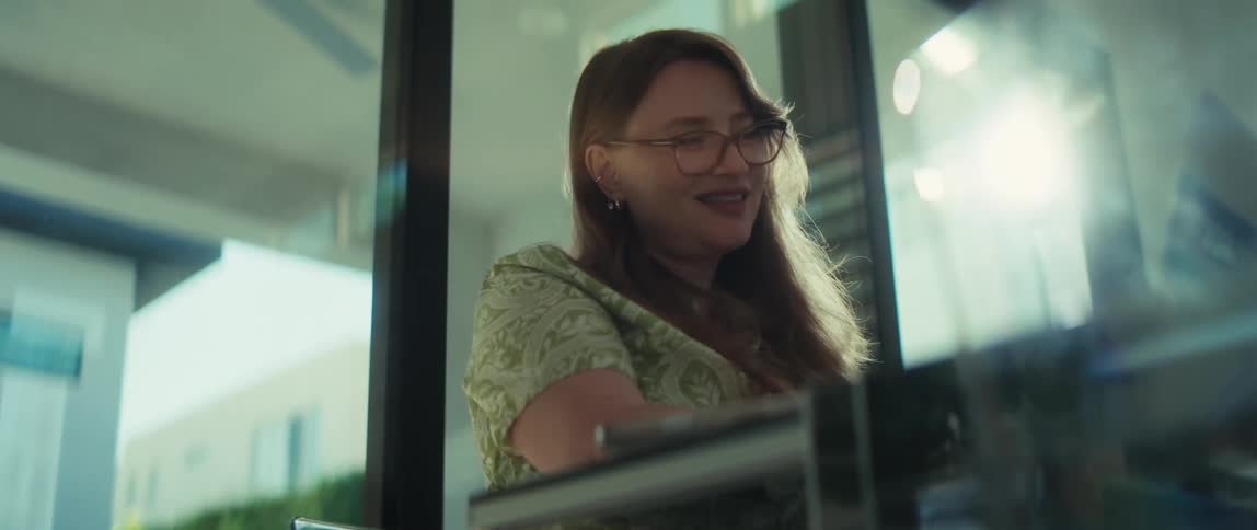 A cheerful young woman with red hair and glasses laughs heartily while working or browsing on her laptop in a sunlit, modern space