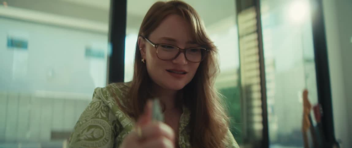 A smiling young businesswoman with red hair and glasses works on her laptop before thoughtfully writing with a pen in a sunlit office