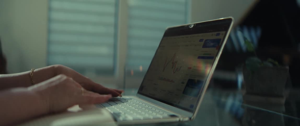 A focused woman wearing glasses types on her laptop while analyzing a financial stock market chart in her home office