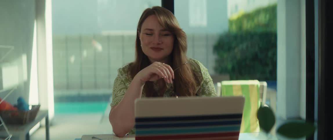 A smiling woman works on her laptop from a sunlit home office, pausing to take off her glasses and rest her eyes