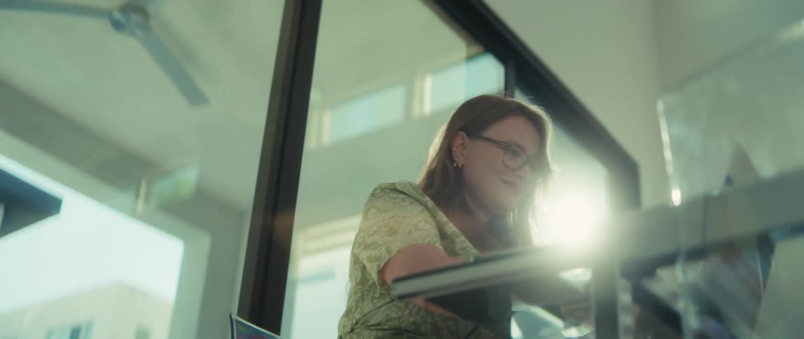 Low-angle shot of a smiling woman in glasses working at her desk as sunlight streams through the window