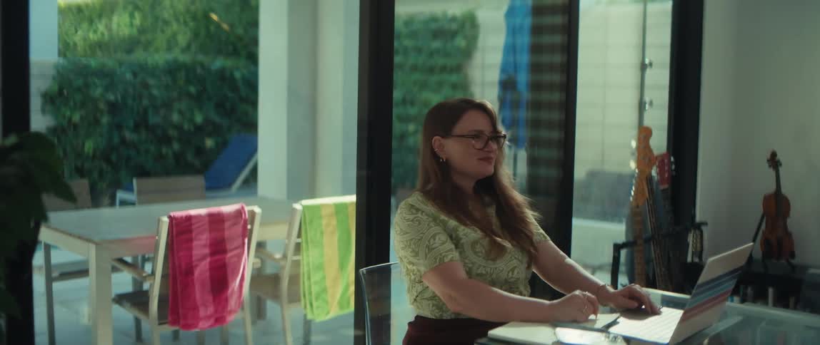 A smiling woman with glasses works on her laptop at a glass table, pausing to think and playfully tap her fingers on the desk in her modern home