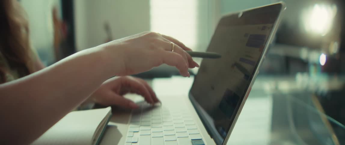A focused woman with glasses works on her laptop in a sunlit room, using a stylus pen to analyze data on the screen