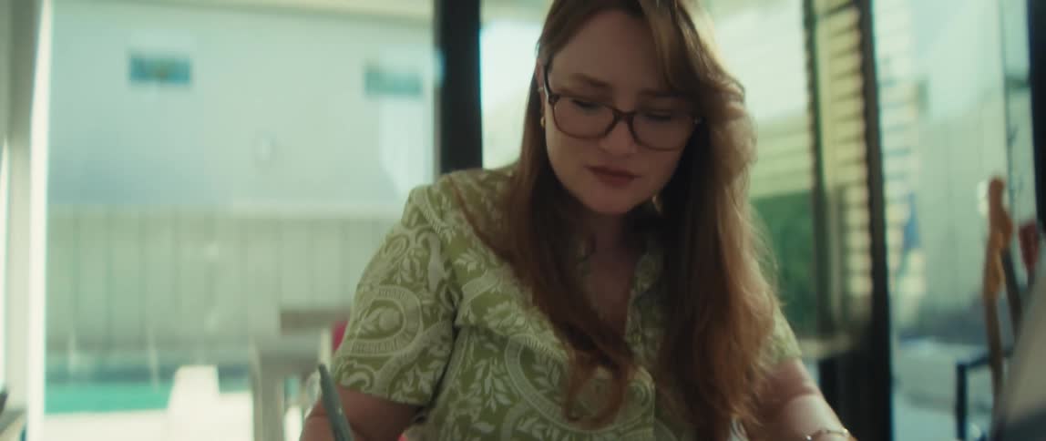 Close-up of a young woman with red hair and glasses writing in a journal at her desk while working from home