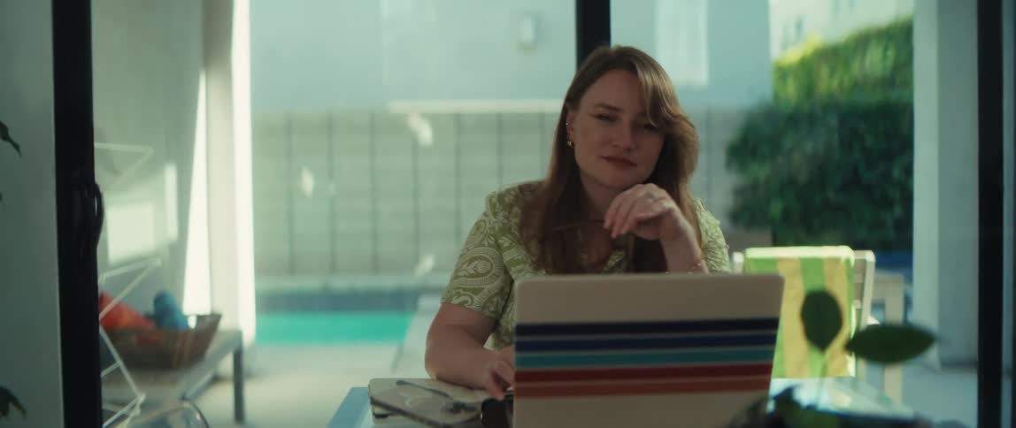 A thoughtful woman removes her glasses and smiles contentedly while working from her sunlit home office with a view of the pool