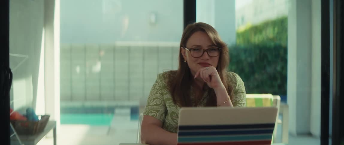 A woman in glasses smiles thoughtfully while working on her laptop in a bright, modern home overlooking a swimming pool