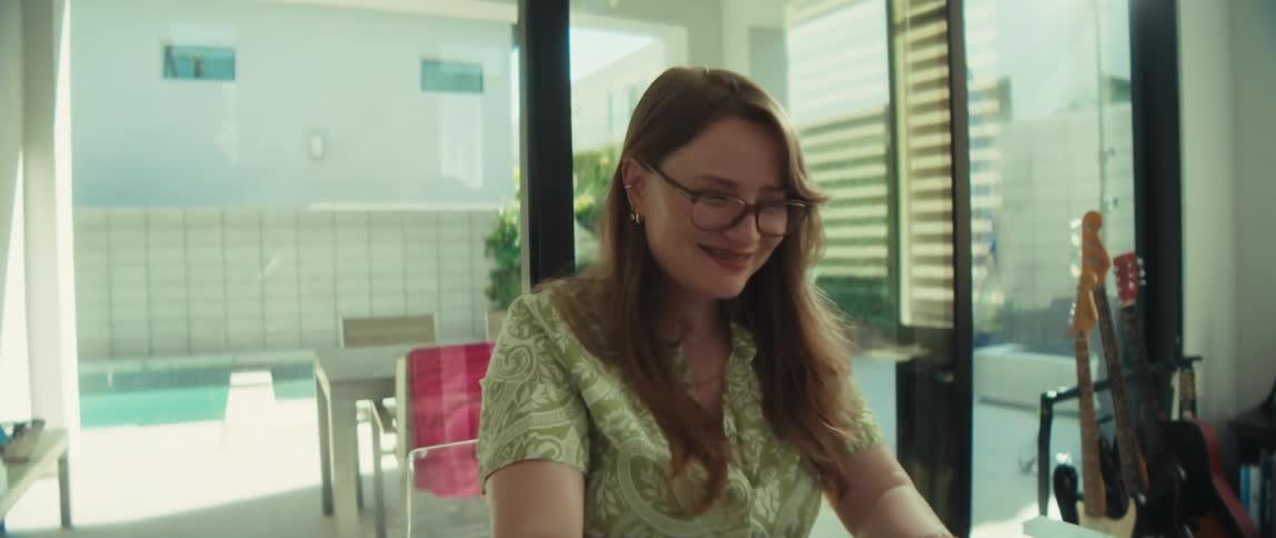 A smiling young woman with glasses works on her laptop in a bright, modern dining room overlooking a backyard pool