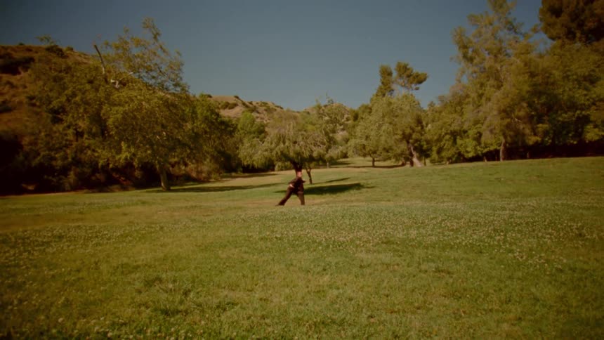 A lone warrior in a dark costume practices with a sword in a sun-drenched grassy field