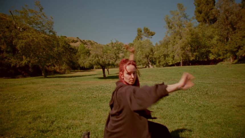 A woman with vibrant red hair performs a slow-motion martial arts routine in a sunlit park, moving with graceful, flowing motions towards the camera