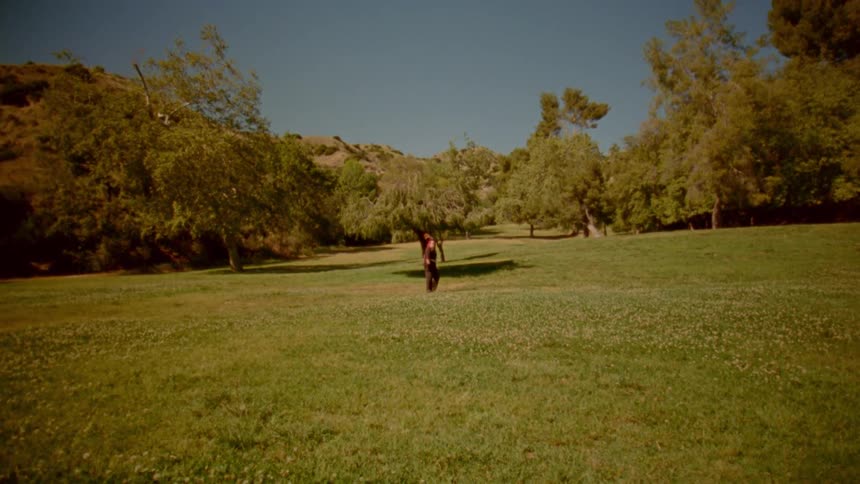 A person in a black and red outfit practices martial arts with a staff in a sunlit grassy meadow, performing slow movements before falling to the ground