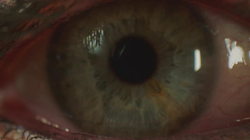 A macro close-up of a human eye with a green iris staring, then blinking to reveal a glittery eyelid