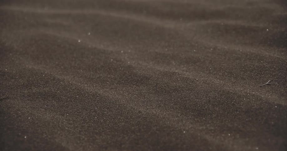 A gentle wind blows fine, dark sand across rippled dunes in a mesmerizing slow-motion close-up
