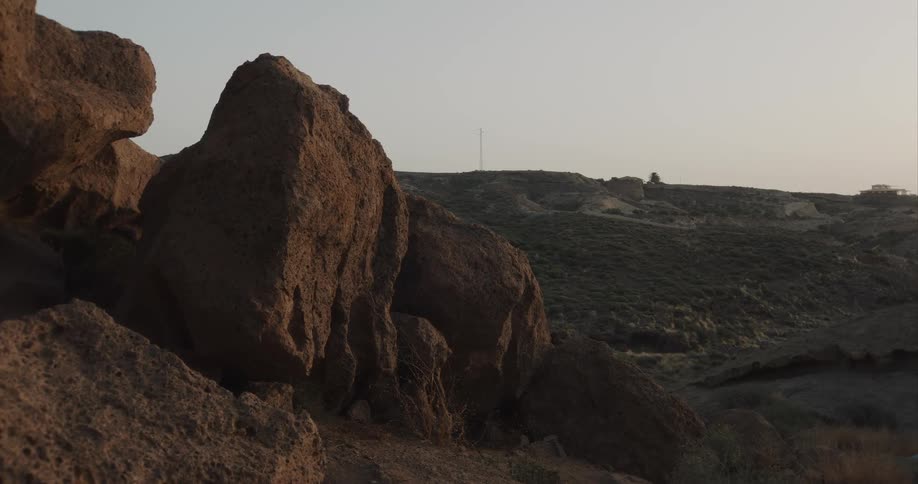 A slow pan reveals a rugged, rocky landscape with green rolling hills in the distance during a hazy sunset
