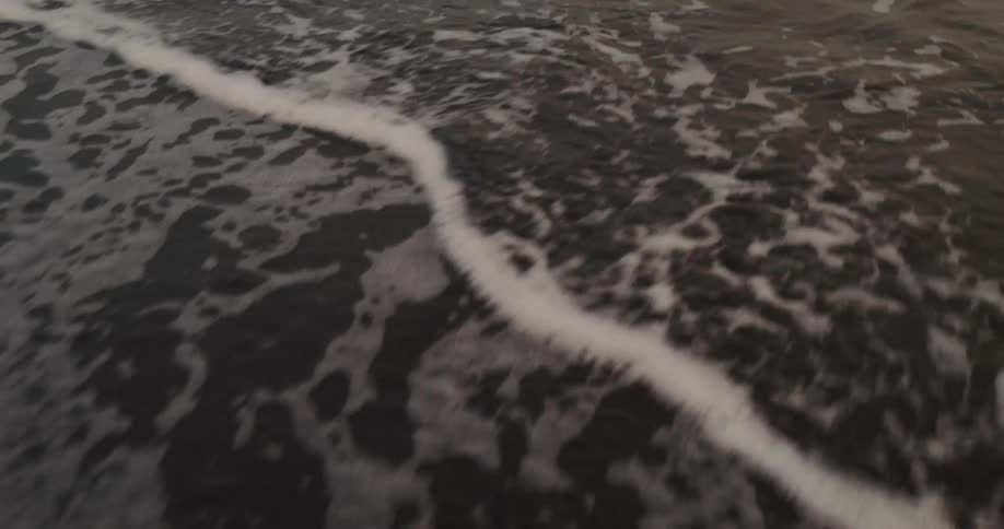 A serene slow-motion shot of gentle ocean waves washing over a dark sandy beach as the tide recedes at dusk