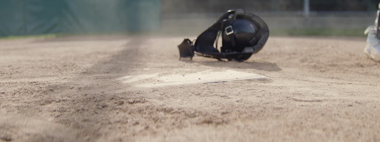 A baseball catcher throws off their mask in slow motion as a player slides safely into home plate, kicking up dust