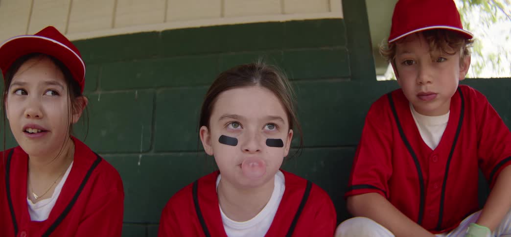 A young girl in a baseball uniform blows a bubble with her gum while sitting in the dugout with her teammates