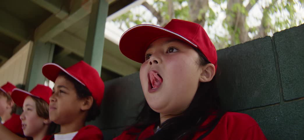 A girl on a little league baseball team blows a bubble with her gum then waves from the dugout bench