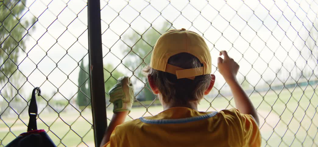A young boy in a yellow baseball uniform watches the game from the dugout through a chain-link fence