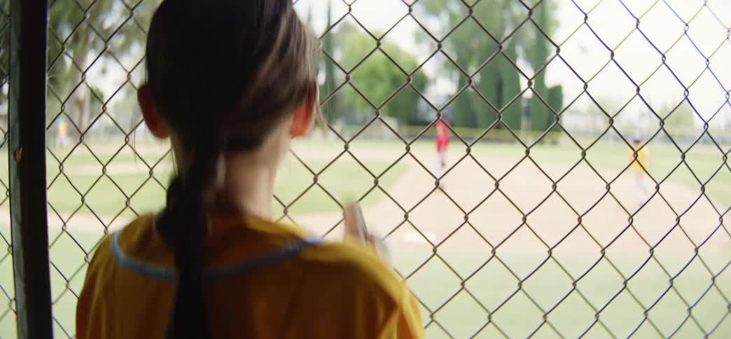A young softball player with a braid waits in the dugout and watches her team play from behind a chain-link fence