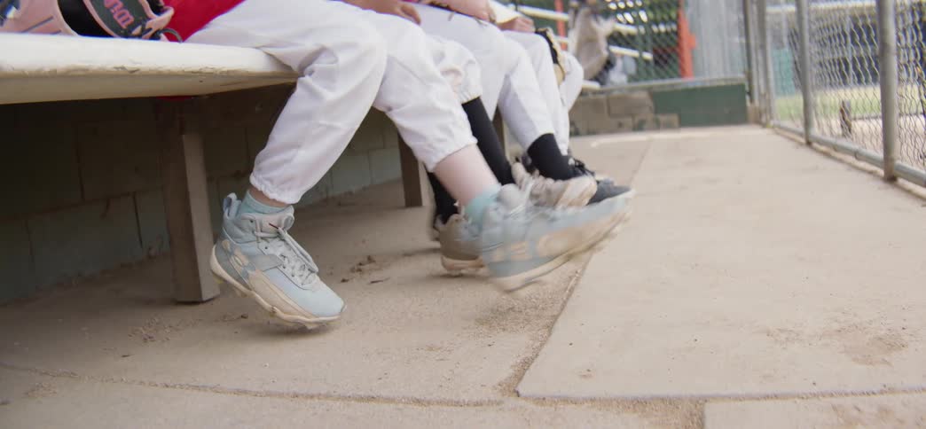 A close-up shot of young baseball players' cleats as they swing their legs back and forth while sitting on the bench in the dugout