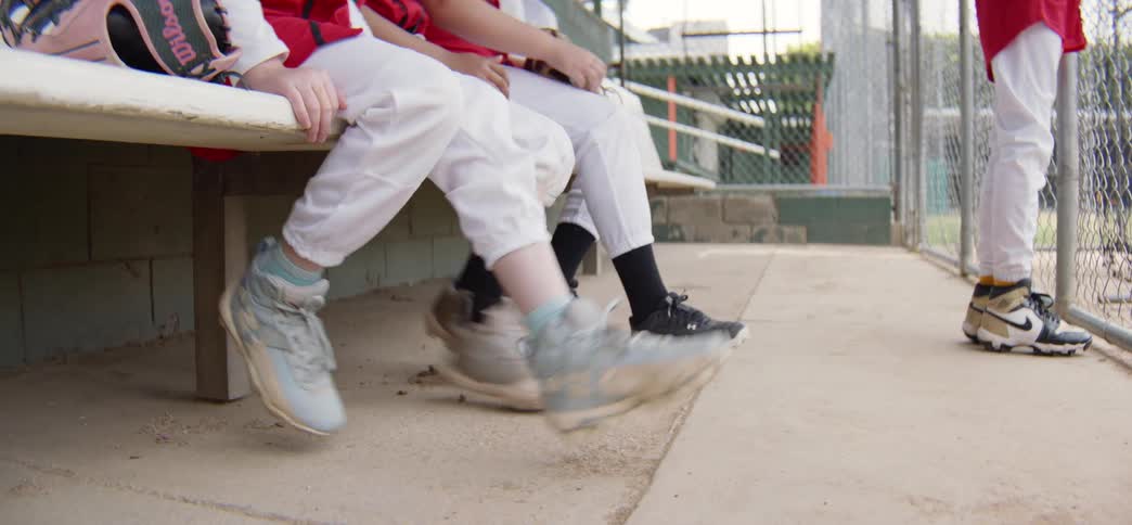 Low-angle shot of youth baseball players swinging their cleated feet while sitting on the bench in a dugout